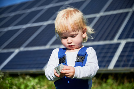 Little child looking at small detail of big solar station. Interested boy learning how to use tools. Cute blond kid holding small piece on background of solar panels at sunny day.の写真素材
