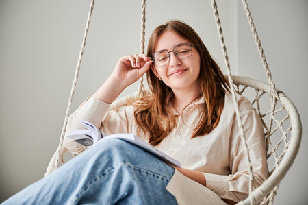 Portrait of smiling girl relaxing with book on indoor swing with closed eyes. Pretty female in glasses excited about story she reading. Cozy afternoon with book in hanging chair in home.の写真素材