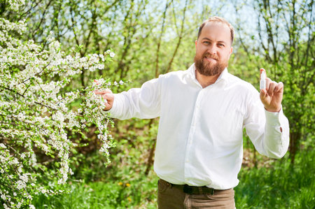 Man allergic using medical nasal drops, suffering from seasonal allergy at spring in blossoming garden. Handsome smiling man showing medicament near blooming tree outdoors. Spring allergy concept.の写真素材