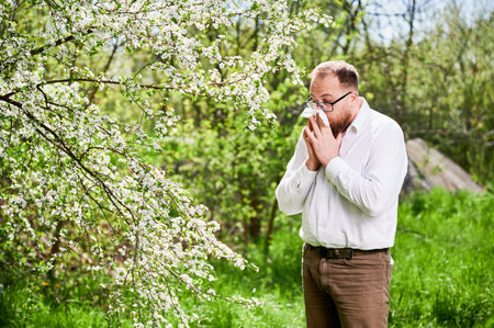 Man allergic suffering from seasonal allergy at spring in blossoming garden at springtime. Young man sneezing and blowing nose with nasal handkerchief in front of blooming tree. Spring allergy conceptの写真素材
