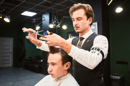 Professional hairdresser in elegant outfit making stylish haircut. Young male client relaxing in chair while getting hairstyle. Handsome barber cutting wet hair.の写真素材
