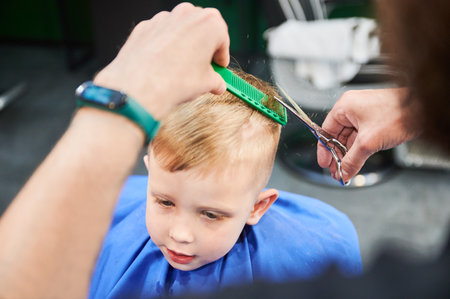 Man barber cutting little boys hair using comb and scissors. Child getting haircut from adult male, likely barber. Professional hairdresser and cute client at modern barbershop.の写真素材