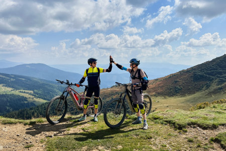 Two cyclists men riding electric bikes outdoors. Male tourists resting on the top of hill, giving high five to each other, enjoying beautiful mountain landscape, wearing helmet and backpack.の写真素材