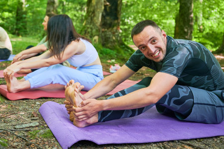 People engaged in stretching exercise outdoors, part of yoga practice. Portrait of happy man stretching forward to reach his toes, sitting on purple yoga mat.の写真素材
