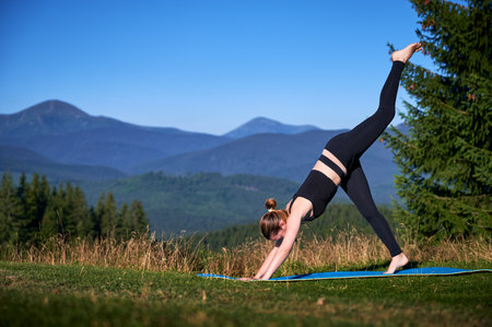 Woman practicing yoga outdoors in the mountains in a serene, natural setting. Female performing yoga pose on mat, with backdrop of beautiful mountain landscape at sunrise or sunset.の写真素材