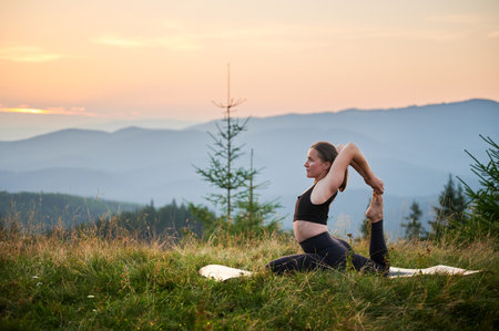 Woman practicing yoga outdoors in the mountains in a serene, natural setting. Female performing yoga pose on mat, with backdrop of beautiful mountain landscape at sunrise or sunset.の写真素材