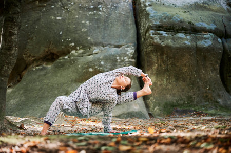 Woman practicing yoga outdoors in forest. Barefoot female on yoga mat surrounded by trees and large rocks, which suggests peaceful, natural environment ideal for meditation or yoga practice.の写真素材