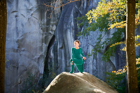 Woman practicing yoga outdoors in forest. Barefoot female on yoga mat surrounded by trees and large rocks, which suggests peaceful, natural environment ideal for meditation or yoga practice.の写真素材