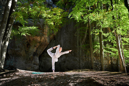 Woman practicing yoga outdoors in forest. Barefoot female on yoga mat surrounded by trees and large rocks, which suggests peaceful, natural environment ideal for meditation or yoga practice.の写真素材