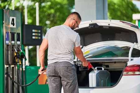 Young man refueling canister with fuel. Skilled driver filling cistern with gasoline in case of unforeseen circumstances. Male adult with pump nozzle refueling canister in car trunk.の写真素材