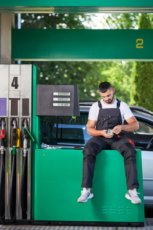 Smiling gas station worker happily counting money. Young operator counting profit while waiting for next customer. Male worker in uniform sitting near gasoline column and holding lot of money.の写真素材