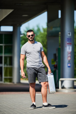 Young man holding canister and waiting for his turn. Smiling male wants to fill canister with gasoline. Handsome man in casual clothes and sunglasses standing on background of gas station.の写真素材