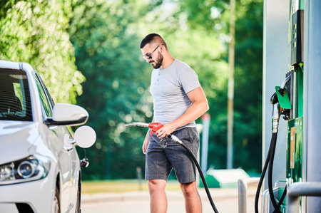 Driver with gasoline pump refilling car gas tank. Confident man refueling his luxury white auto. Man in casual clothes and sunglasses at modern gas station.の写真素材
