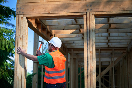 Carpenter constructing wooden frame house near the forest. Bearded man hammering nails into structure while wearing protective helmet and construction vest. Concept of modern ecological construction.の写真素材