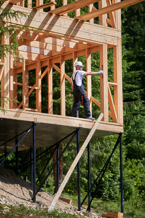 Carpenter building two-story wooden frame home near the forest. Bearded man confirms wall evenness with level while donning protective gear and helmet. Concept of modern and eco-friendly building.の写真素材