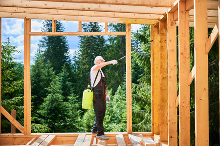 Laborer constructing wooden frame house near forest. Man treating woods, applying fire retardant using sprayer, while dressed in protective suit, helmet. Concept of modern eco-friendly construction.の写真素材