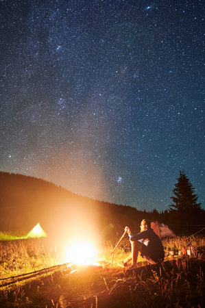 Night camping in mountains under starry sky. Man sitting near campfire, holding stick. Male hiking, traveling, admiring fire, landscape, resting, enjoying. Concept of harmony with nature.の写真素材