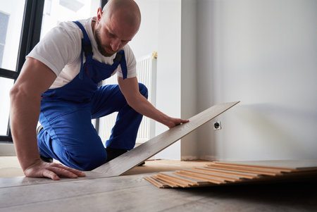 Bearded man in work overalls installing laminate timber flooring in apartment under renovation. Male construction worker laying laminate wood plank on floor underlayment in living room.の写真素材