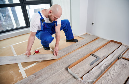 Man in work overalls laying laminate timber planks on the floor in apartment under renovation. Male worker installing laminate wooden flooring in flat. Home renovation and wood flooring concept.の写真素材