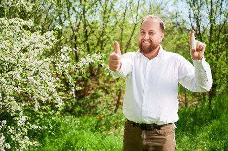 Man allergic using medical nasal drops, suffering from seasonal allergy at spring in blossoming garden. Handsome smiling man showing thumbs up near blooming tree outdoors. Spring allergy concept.の写真素材