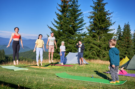 Group of people doing yoga pose outdoor in camping in the mountains. Adults and children standing on yoga mats, each doing a yoga pose under a clear blue sky in the morning. Young boy is instructor.の写真素材