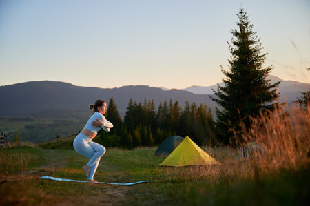 Woman practicing yoga outdoors in the mountains in a serene, natural setting near camping. Female performing yoga pose on mat, with backdrop of beautiful mountain landscape at sunrise or sunset.の写真素材