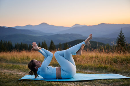 Woman practicing yoga outdoors in the mountains in a serene, natural setting. Female performing yoga pose on blue mat, with backdrop of beautiful mountain landscape at sunrise or sunset.の写真素材