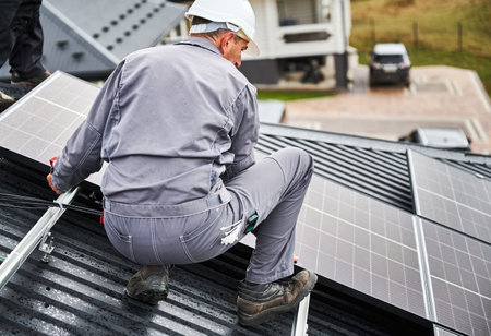 Builder mounting photovoltaic solar modules on roof of house. Back view of man engineer in helmet installing solar panel system outdoors. Concept of alternative and renewable energy.の写真素材