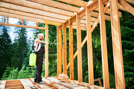 Laborer constructing wooden frame house near forest. Man treating woods, applying fire retardant using sprayer, while dressed in protective suit, helmet. Concept of modern eco-friendly construction.の写真素材