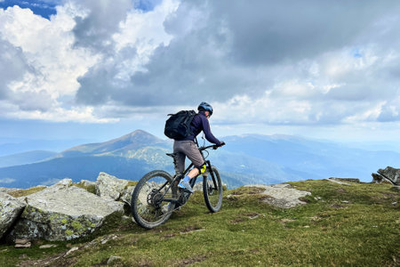 Cyclist man riding electric mountain bike outdoors. Male tourist biking along grassy trail in the mountains, wearing helmet and backpack. Concept of sport, active leisure and nature.の写真素材