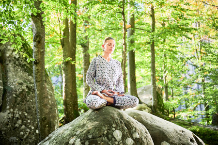 Woman practicing yoga outdoors in forest. Barefoot female on yoga mat surrounded by trees and large rocks, which suggests peaceful, natural environment ideal for meditation or yoga practice.の写真素材