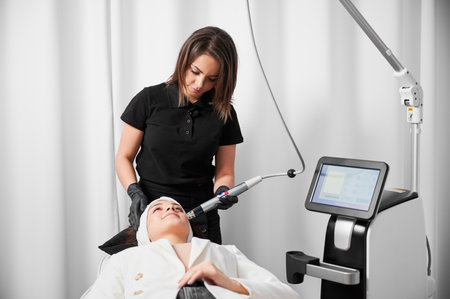 Female cosmetologist using radiofrequency microneedling device while performing face lifting procedure. Young woman lying near cosmetology equipment and receiving skincare treatment.の写真素材