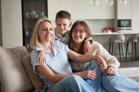 Lovely family relaxing on couch in living room. Little boy hugging his sister and mom. Cozy spending time with family indoors. In background, modern kitchen, neutral colors, minimalistic design.の写真素材