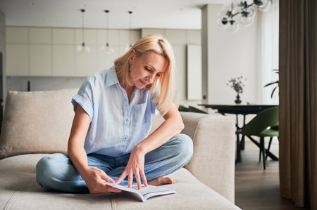 Woman sitting comfortably on couch, deeply engaged in reading book. Cozy living space with modern aesthetic on background. Person enjoying quiet moment of solitude, immersed in reading.の写真素材
