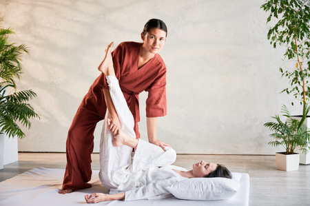 Masseuse making thai yoga massage. Woman therapist doing traditional massage treatment, stretching female patients body. Client lying on mat, enjoying hand massage in calm atmosphere.の写真素材