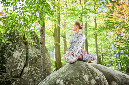 Woman practicing yoga outdoors in forest. Barefoot female on yoga mat surrounded by trees and large rocks, which suggests peaceful, natural environment ideal for meditation or yoga practice.の写真素材
