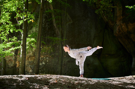 Woman practicing yoga outdoors in forest. Barefoot female on yoga mat surrounded by trees and large rocks, which suggests peaceful, natural environment ideal for meditation or yoga practice.の写真素材