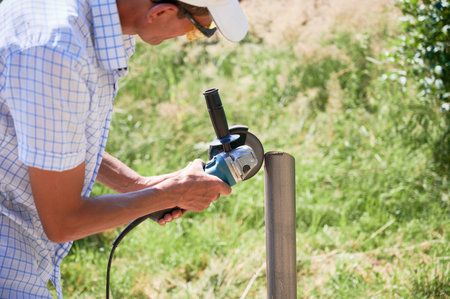 Male worker cutting metal pole with electric circular saw. Man in protective glasses using hand-held grinder while preparing aluminum pole for solar panel installation.の写真素材