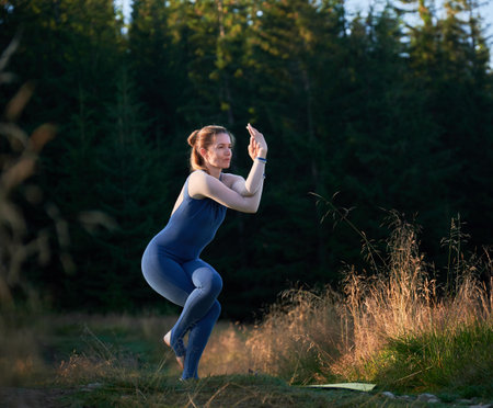 Middle-aged woman doing eagle pose. Young female practicing yoga poses in coniferous forest. Woman in dark blue tracksuit exercising in morning.の写真素材