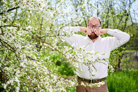 Man allergic suffering from seasonal allergy at spring in blossoming garden at springtime. Young bearded man with glasses having runny nose, closing nose by hands in front of blooming tree.の写真素材