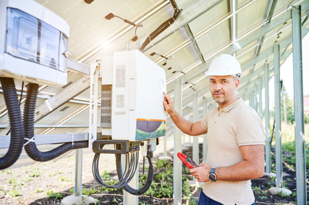 Professional worker checking voltage on solar inverter. Male using current probe to measure output voltage. Man in helmet pointing at inveter dial on back side of PV panel, looking to the camera.の写真素材