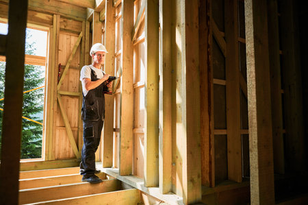 Carpenter constructing wooden framed house. Man worker working with screwdriver, wearing work overalls and helmet. Concept of modern eco-friendly construction.の写真素材