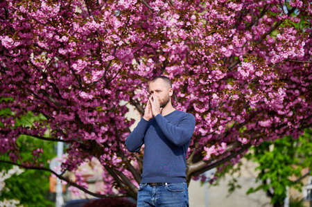 Man allergic suffering from seasonal allergy at spring in blossoming garden at springtime. Bearded young man sneezing and having runny nose in front of blooming tree. Spring allergy concept.の写真素材