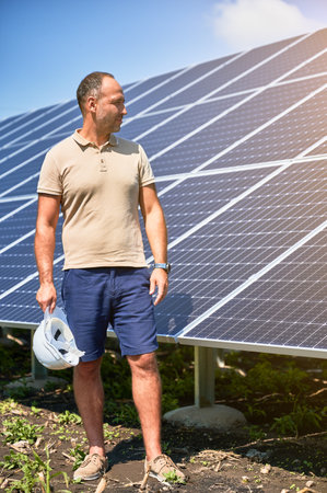 Professional worker glad about work he did. Installed PV panel ready to work. Adult male looking at solar panel with helmet in hand.の写真素材