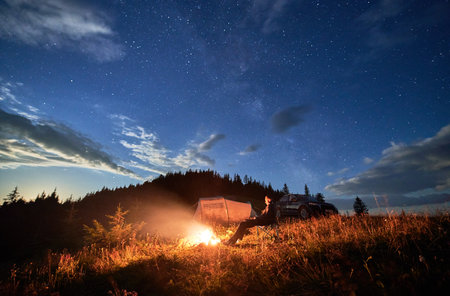 Camping in mountains under evening cloudy sky. Young man having a rest next to campfire, near tourist tent and off-road car on hill. Travelling man sitting and stargazing at night.の写真素材