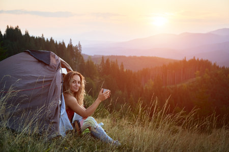 Woman traveller camping outdoors at sunset. Beautiful female sitting in tents entrance on grass with cup of tea. Young tourist hiking, having rest in campsite. Concept of tourism in mountains.の写真素材