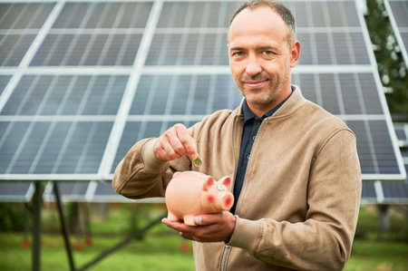 Man making investment in alternative ecological resources. Male putting his money in piggy bank for future profit. Investor in casual clothes holding moneybox on background of solar panel.の写真素材