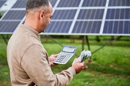Confident investor counting profit he got from investing in green energy. Glad man holding huge sum received by successfull investment. Back view of man with calculator and money next to PV battery.の写真素材