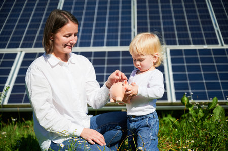 Modern mom and her little kid saving money in piggy bank. Mother teaching son how to invest money in future. Smiling mom and her kid putting savings into piggybank on background of solar panels.の写真素材