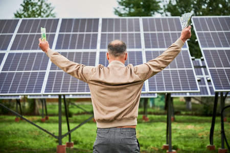 Back view of man happy about profit he got from investing in green energy. Man exulting with huge sum in hands and standing next to solar battery he bought.の写真素材
